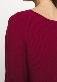 Femme portant un pull côtelé rouge foncé à large encolure et une boucle d'oreille en perle pendante sur un fond blanc uni.