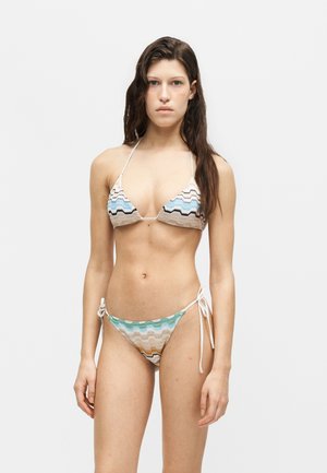 Žena s dlhými hnedými vlasmi v viacfarebnom trojuholníkovom bikinách so vzorom cik-cak, stojaca na bielom jednoduchom pozadí.