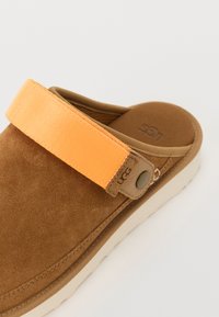 Bruine nubuck slip-on sandaal met een heldere oranje band. Heeft een gevoerde binnenzool en een dikke, witte rubberen buitenzool. Logo aan de zijkant.