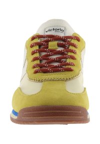 Vista frontale di una sneaker gialla e crema con lacci marroni e suola marrone con dettagli a strisce blu e bianche.