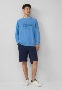 Blauwe sweatshirt met zwart S.Oliver-logo, gecombineerd met donkerblauwe shorts en witte sneakers. Glad materiaal, casual ontwerp, relaxte pasvorm.