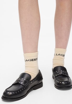 Mocassins en cuir noir avec des accents cloutés ronds et une finition lisse, portés avec des chaussettes à crème portant un texte de marque noir.