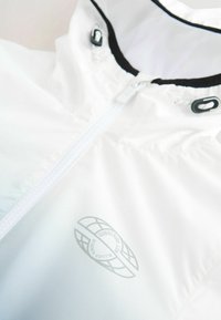 Veste légère blanche avec fermeture éclair, accents noirs autour du col et un logo subtil imprimé sur le tissu. Texture lisse.