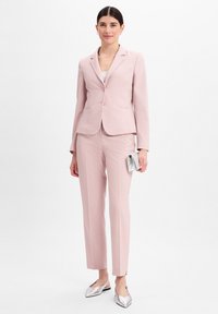 Lichtroze op maat gemaakt kostuum met een getailleerde blazer en rechte broek, met een gestructureerde stof, gecombineerd met zilveren flats en een kleine zilveren clutch.