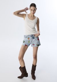 Top de tirantes color beige claro con escote redondo, combinado con pantalones cortos de denim con bordado colorido de frutas, usado con botas marrones de tobillo holgadas.