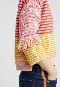 Frau trägt einen mehrfarbigen, gestreiften Strickpullover mit Fransen an den Ärmeln und blaue Jeans mit einem kupferfarbenen Seitenstreifen.