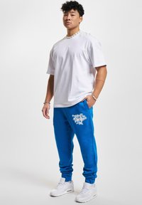 Witte t-shirt met korte mouwen gecombineerd met heldere blauwe trainingsbroek met een graffiti-stijl wit logo. Draag het met witte sportschoenen.