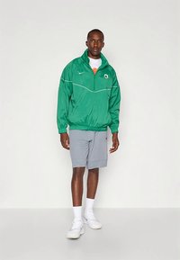 Grüne Windjacke mit halbem Reißverschluss, elastischen Bündchen und Logo; kombiniert mit hellgrauen Shorts und weißen Turnschuhen vor einfarbigem Hintergrund.