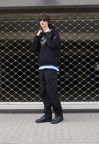 Schwarzer Hoodie mit grafischem Druck, über einem hellblauen Hemd getragen. Schwarze Cargo-Hosen und schwarze Sneakers runden den Look ab. Gewebter Mesh-Hintergrund.