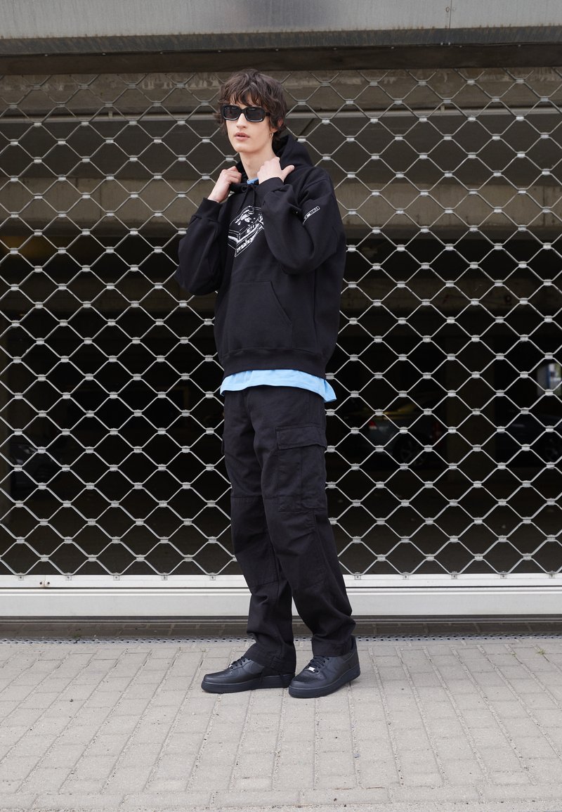 Schwarzer Hoodie mit grafischem Druck, über einem hellblauen Hemd getragen. Schwarze Cargo-Hosen und schwarze Sneakers runden den Look ab. Gewebter Mesh-Hintergrund.