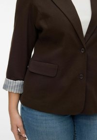 Blazer marron avec une poche avant unique, deux boutons et des poignets rayés retroussés. Associé à un jean denim bleu. Texture lisse.