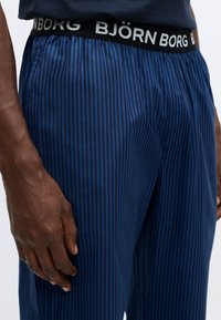 Plave i crne prugaste pidžama hlače s elastičnim pojasom na kojem se nalazi bijeli logo "BJÖRN BORG". Materijal izgleda glatko i lagano.
