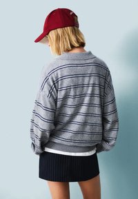 Grijs gestreepte trui met blauwe lijnen, ronde hals, en een losse pasvorm, gecombineerd met een zwarte pinstripe rok en een rode pet. Aanzicht van achter.