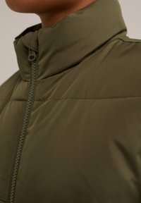 Olivgrön vadderad jacka med hög krage, med en central dragkedja och slät textur. Ren, minimalistisk design utan synliga mönster.