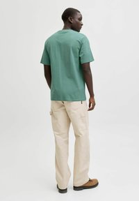 Roheline lühikeste varrukatega puuvillane t-särk, kombineeritud beežide taskutega cargo-pükstega, kantud pruunide jalanõudega. Taust on neutraalne.