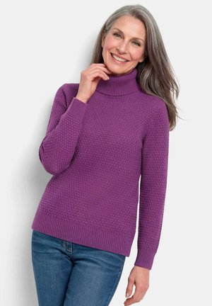 Femme mûre souriante aux longs cheveux gris portant un pull à col roulé violet texturé et un jean bleu, posant devant un fond blanc.