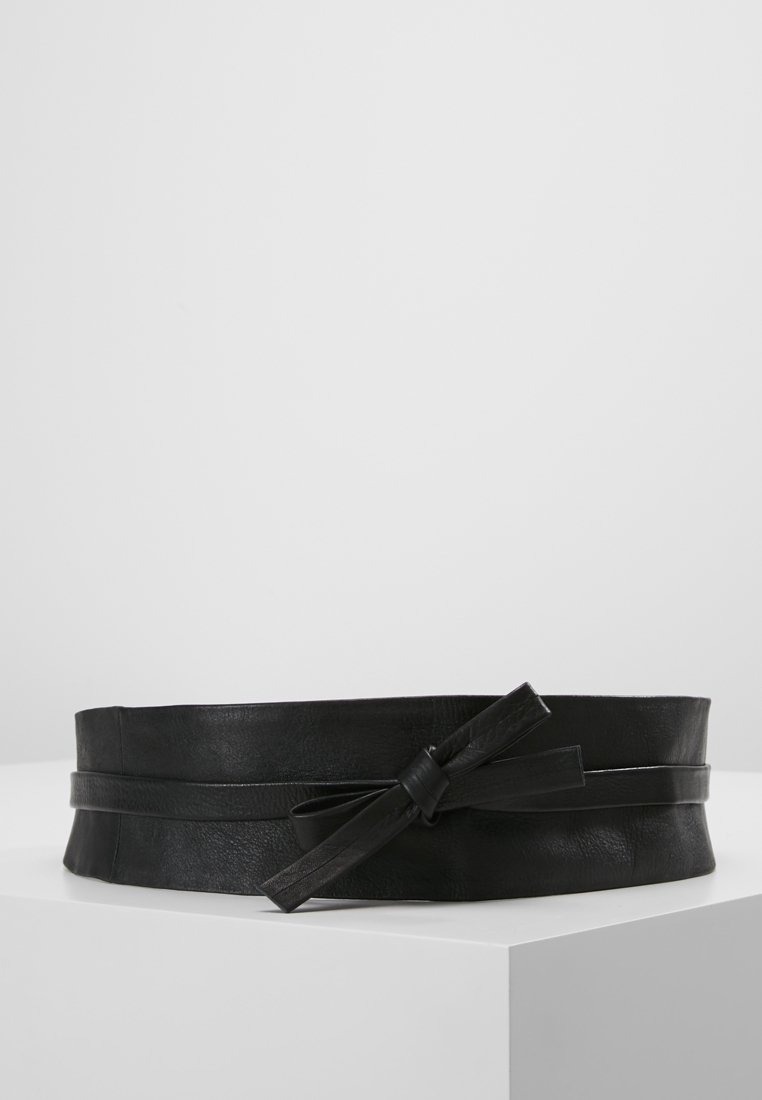 Ceinture large en cuir noir avec une fine lanière en cuir nouée en nœud à l'avant, reposant sur une surface blanche.