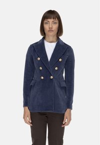 Giacca doppiopetto blu navy in tessuto morbido, con bottoni dorati, colletto a punte e tasche laterali, indossata sopra una camicia bianca.