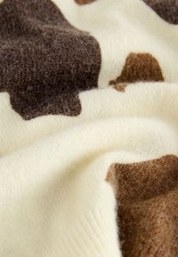 Pleteni pulover iz mehke volne; ima kremasto osnovo z nepravilnimi rjavimi madeži, ki prikazujejo teksturirano površino in gladko padajoč material.