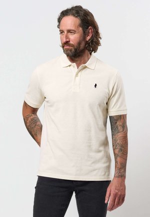 Hombre con cabello castaño y barba, vistiendo una camiseta polo crema y pantalones negros, mostrando brazos tatuados, de pie frente a un fondo blanco liso.