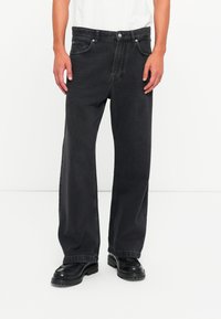 Mand iført løse sorte jeans med knap og lynlås, parret med sorte lædersko foran en almindelig hvid baggrund.