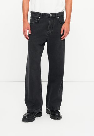 Mann iført løse sorte jeans med knapp og glidelås, kombinert med sorte lærsko mot en enkel hvit bakgrunn.