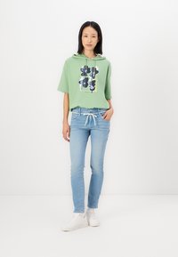 Groene hoodie met bloemenprint, gecombineerd met lichtblauwe spijkerbroek en witte sneakers. De stof lijkt zacht en heeft een ontspannen pasvorm.