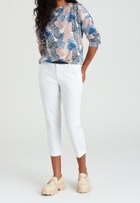 Blusa stampata con motivi astratti in blu, rosa e nero, abbinata a pantaloni bianchi a 3/4 e mocassini beige chunky. Tessuto morbido, vestibilità rilassata.