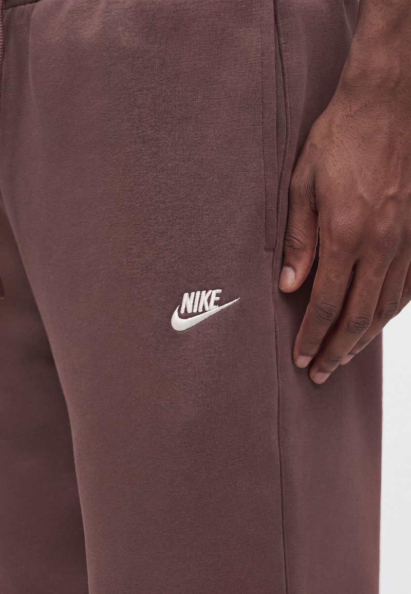 Nahaufnahme einer dunkelbraunen Nike Jogginghose mit weiß gesticktem Logo in der Nähe der Tasche und der Hand einer Person, die daneben ruht.