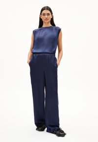 Navy-satin Top mit ärmellosem Design, kombiniert mit locker sitzenden navy-blauen Satin-Hosen. Schwarze Schuhe mit Fransenverzierung runden den Look ab.