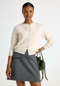 Donna che indossa un cardigan beige con bottoni e una gonna a quadri grigia, mentre tiene una piccola borsa verde con una mano sulla vita.