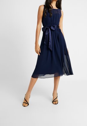 Woman wearing a sleeveless navy midi dress with pleats and a satin bow belt, paired with black strappy heeled sandals, on white background.