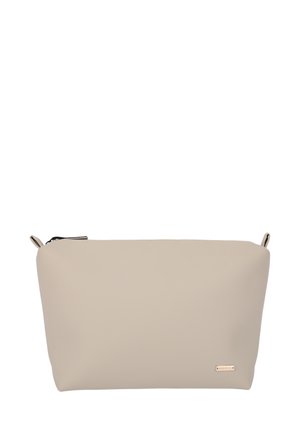Kvinde med krøllet hår iført beige rullekrave og bukser holder en beige clutch mod en hvid baggrund.