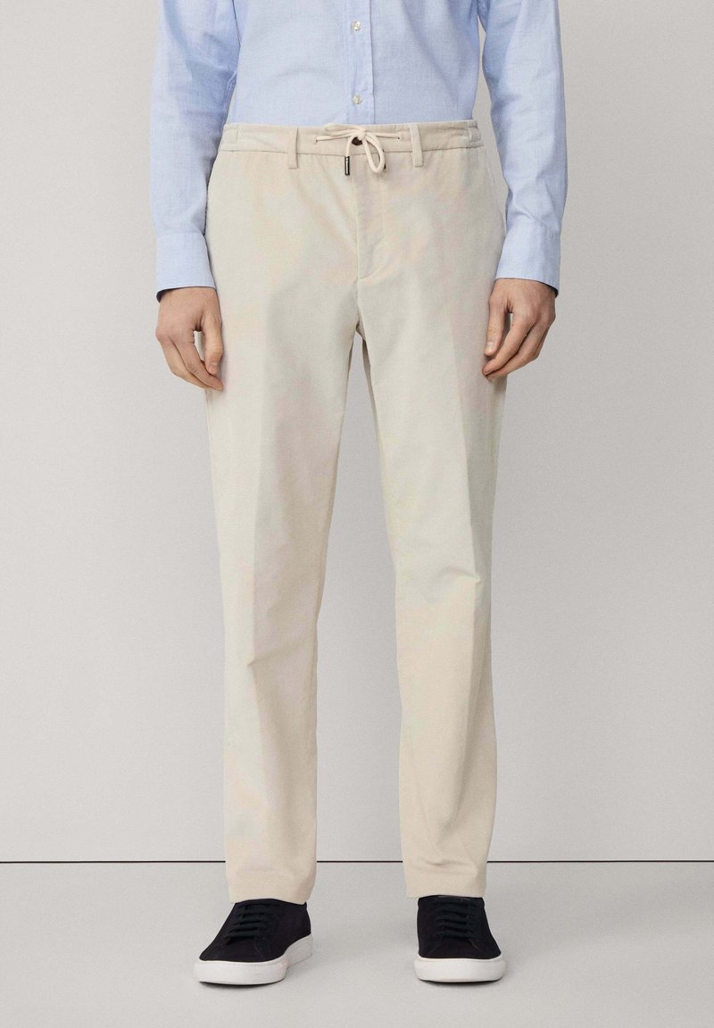Pantalones de algodón beige con cintura ajustable, perneras rectas y textura mínima, combinados con zapatillas oscuras y una camisa de botones azul claro.