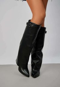 Bottes en cuir noires, montantes jusqu'au genou, avec un design flou, un bout pointu et un détail de bracelet à boucle. Texture lisse avec une finition brillante.
