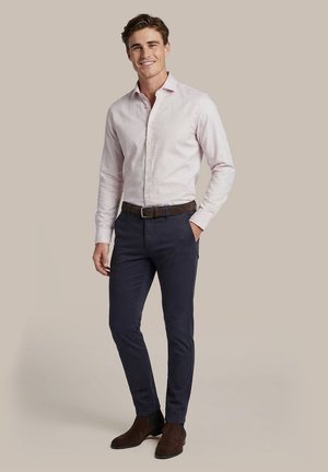 Jonge man glimlacht, draagt een lichtroze overhemd met knopen, donkerblauwe broek, bruine riem en bruine suède laarzen, staand voor een beige achtergrond.
