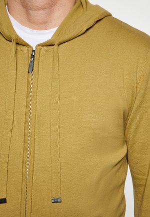 Olijfgroene zip-up hoodie gemaakt van ribbelstof. Beschikt over een trekkoordcapuchon, zilveren rits en een textuurovervlak voor extra detail.