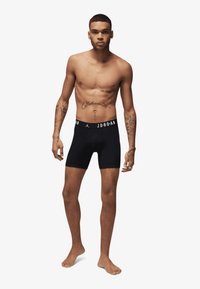 Sorte boxershorts med en mærket elastisk talje, med et tætsiddende design. Modellen står barfodet, med eksponeret overkrop, der viser tatoveringer.
