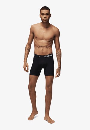 Svarte boxer-shorts med en merket elastisk midjebånd, med en tettsittende design. Modellen står barføtt, med eksponert overkropp, og viser frem tatoveringer.