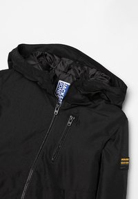 Czarna kurtka z kapturem, z zamkami z przodu i na klatce piersiowej, pikowana podszewka oraz mała żółto-czarna naszywka marki na górnej części rękawa.