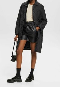 Sort quiltet frakke med krave, beige top, sorte lædershorts, sorte strømper og blanke sorte sko. Enkel håndtaske i hånden.
