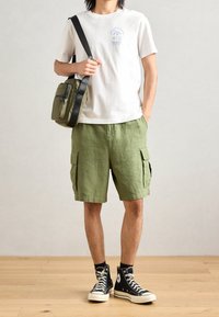 Bílé bavlněné tričko s modrou grafikou, zelené cargo šortky, černé vysoké tenisky a zelená crossbody taška se zipovými detaily.
