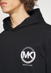 Black cotton hoodie featuring a large circular logo in silver and white with "MICHAEL KORS" text underneath. Soft texture, casual design.