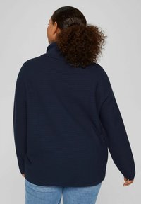 Maglione strutturato blu navy con colletto alto, maniche lunghe e vestibilità ampia. Indossato con jeans azzurri, visto da dietro.