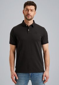 Zwart poloshirt van katoen met een klassieke kraag, twee knopen en korte mouwen. Bevat een klein patch op de borst en accenten aan de zijkanten.