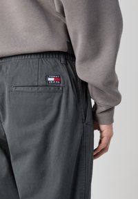 Harmaat kangashousut, joissa on elastinen vyötärö, sivutaskut ja takatasku, jossa on punainen, valkoinen ja sininen "TOMMY JEANS" -logo.