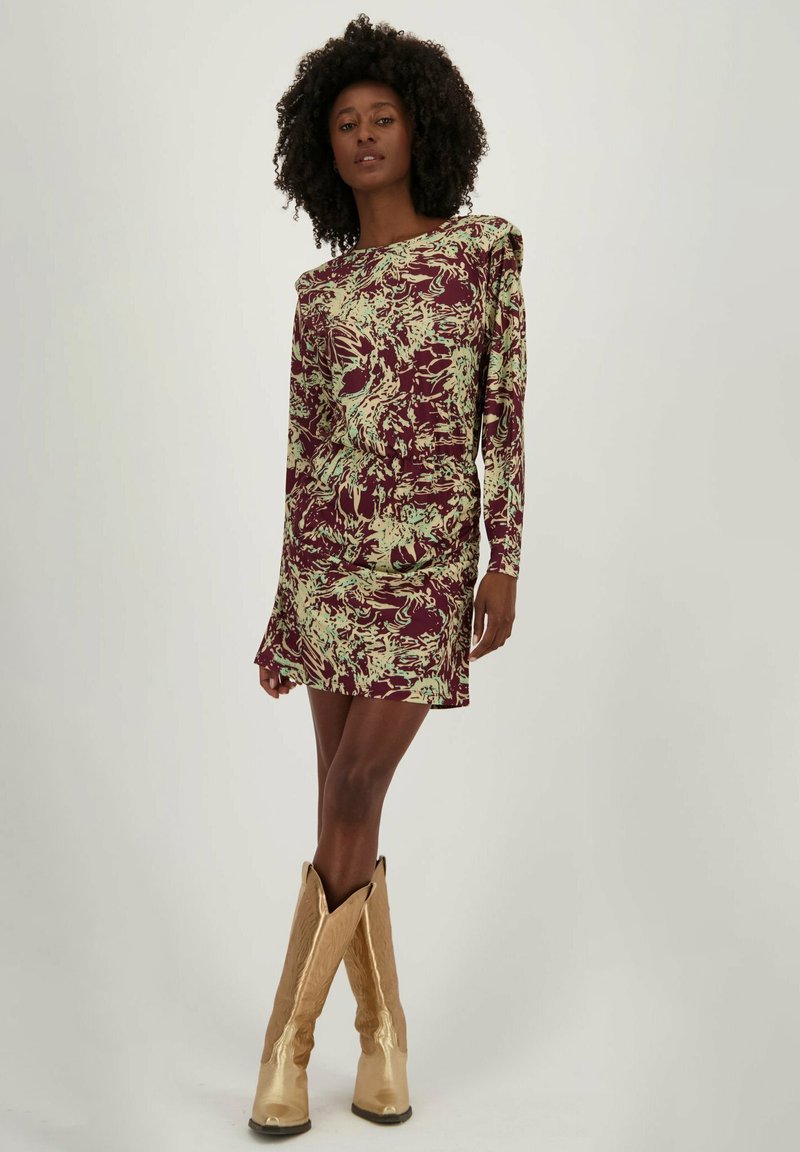 Langemouwen jurk in bordeauxrood met een groen abstract patroon, met een getailleerde taille en een lichte drape. Gekweld met glanzende gouden kniehoge laarzen.