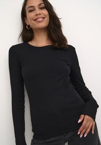 Crni dugih rukava top s rebrastom teksturom i okruglim izrezom. Ima prilagođene rukave i suptilne šavove. Nosi se s tamnosivim trapericama.