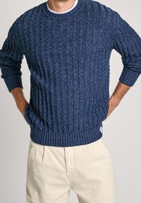 Marineblauer Strickpullover mit einem Zopfmuster, geripptem Rundhalsausschnitt und Bündchen. Kombiniert mit hellen Hosen. Strukturiertes Gewebe.