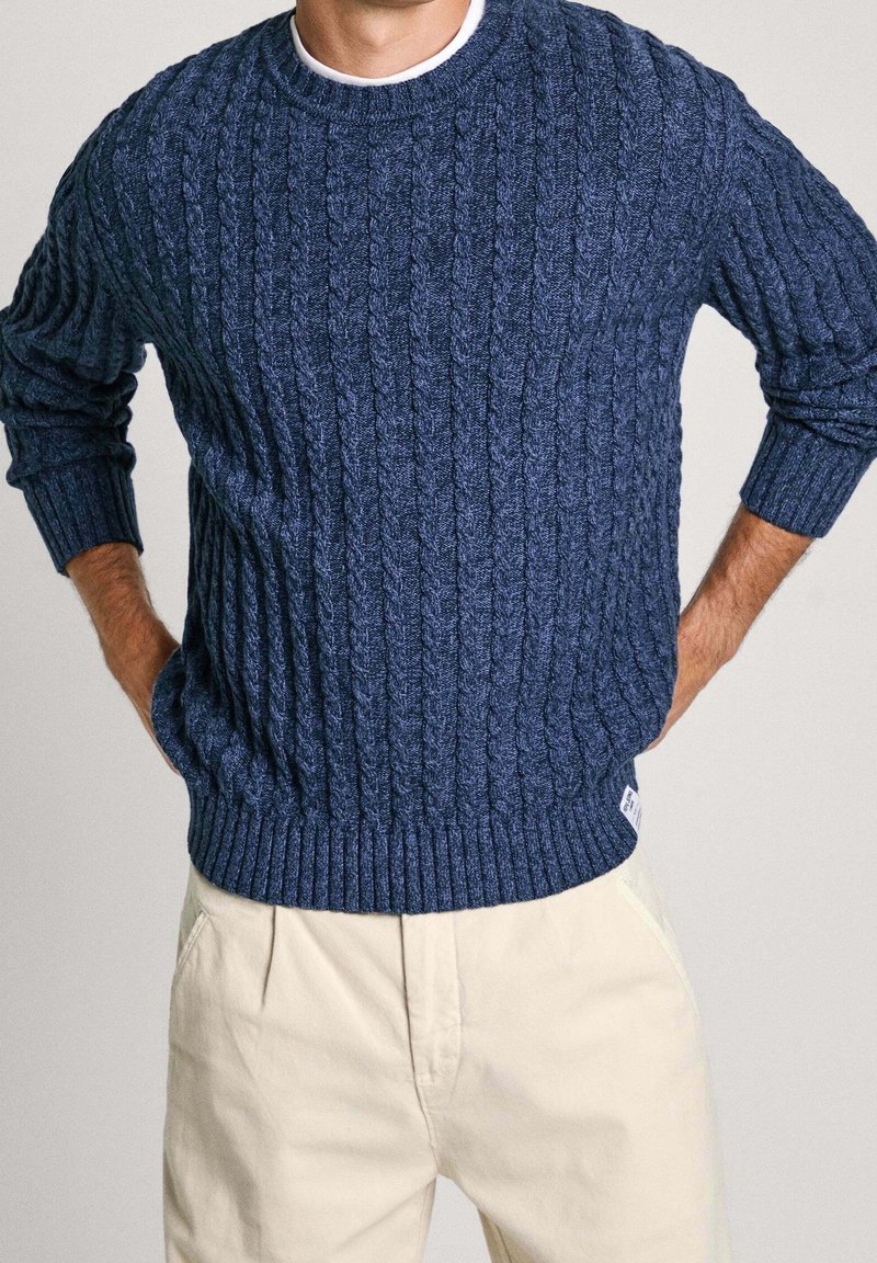 Marineblauer Strickpullover mit einem Zopfmuster, geripptem Rundhalsausschnitt und Bündchen. Kombiniert mit hellen Hosen. Strukturiertes Gewebe.